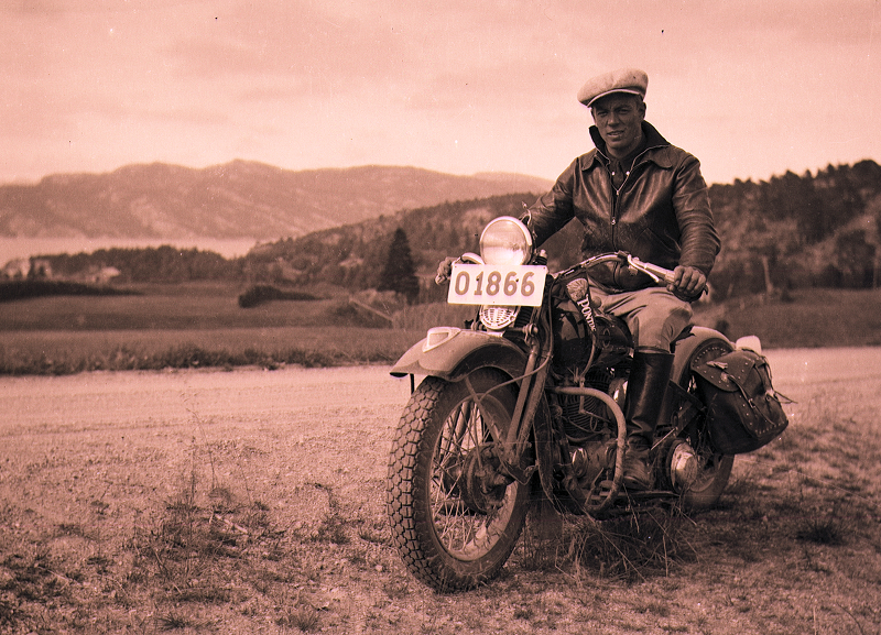 Vintage photo of Gustaf Stenmark seated on a classic motorcycle with saddlebags, parked overlooking hills and water in the distance