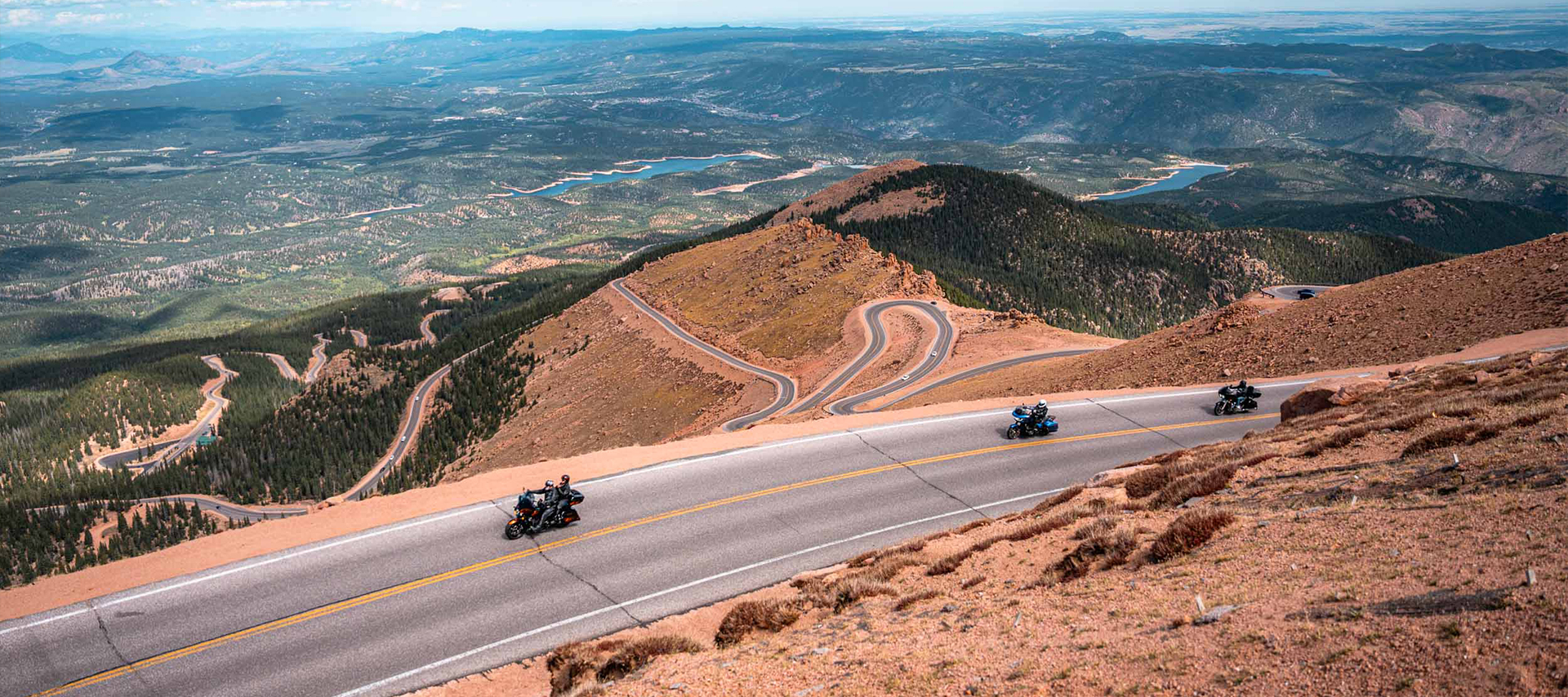 A wide shot looking down of three Harley-Davidson motorcycles ascending Pikes Peak, with the road winding in the distance and landscape extending to the horizon.