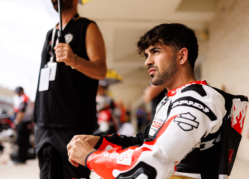 Racer in full gear sits in pit lane and looks into the distance while crew members and equipment line the background