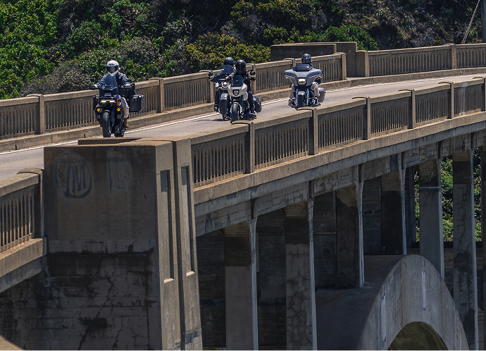 Three riders travel together across Bixby Bridge with repeating concrete arches and guardrails above water