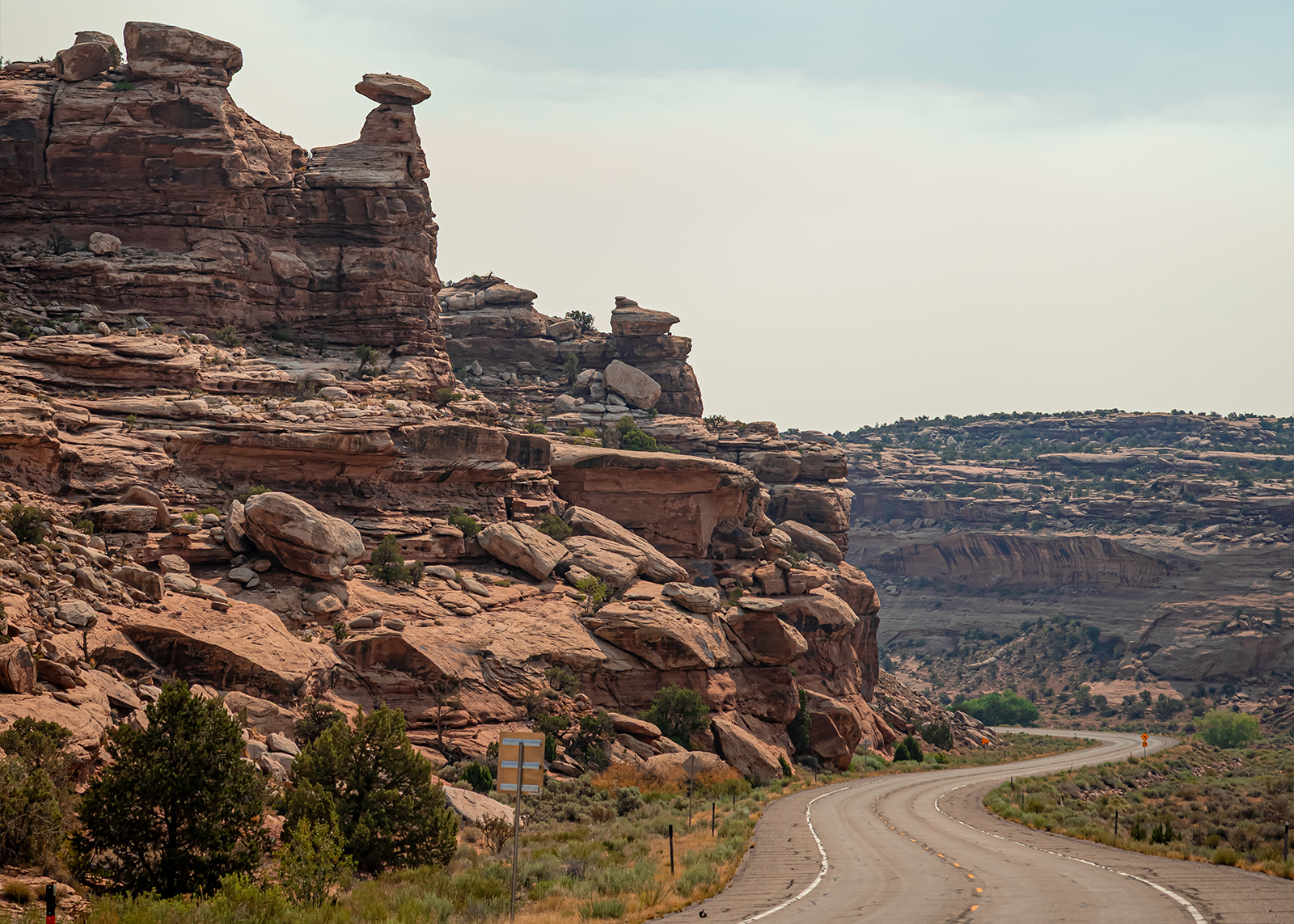 Winding desert highway curves past dramatic red rock formations under a hazy sky in the canyon.