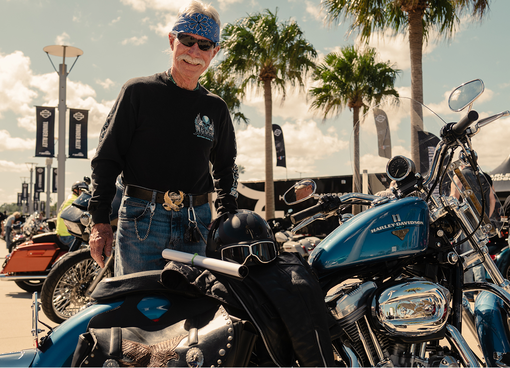 Rider standing beside a blue touring motorcycle among rows of bikes and palm trees at a rally