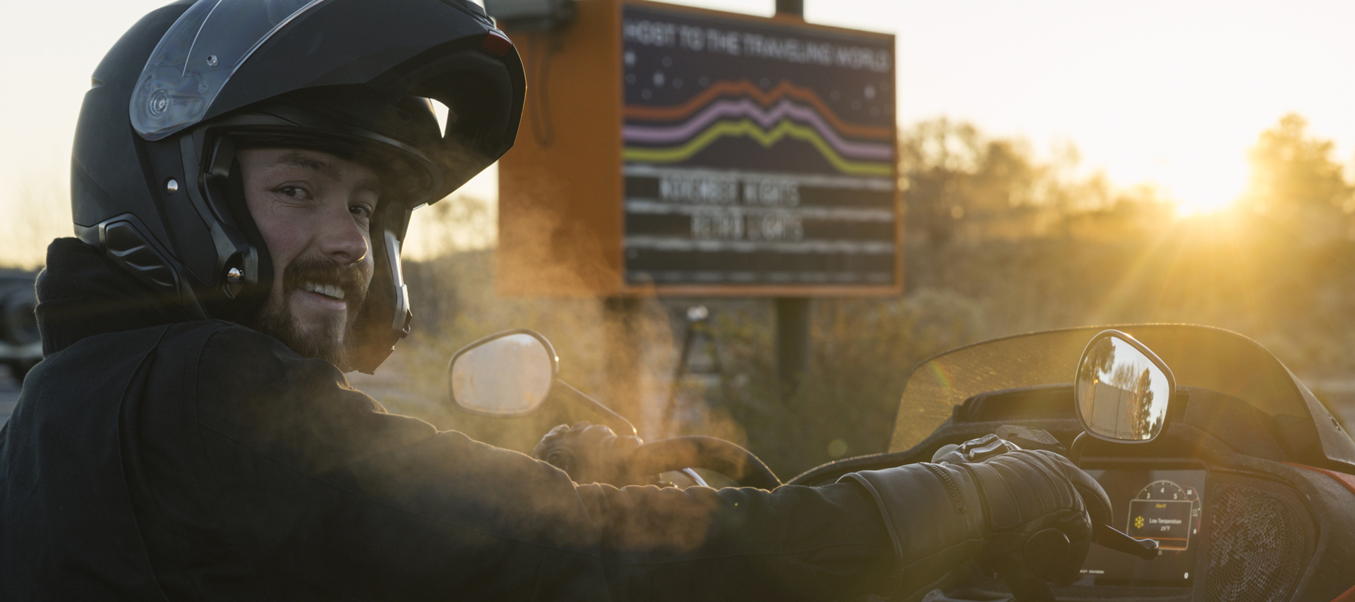 A motorcyclist wearing a helmet and gloves rides at sunset, with cold fog from their breath visible in the air