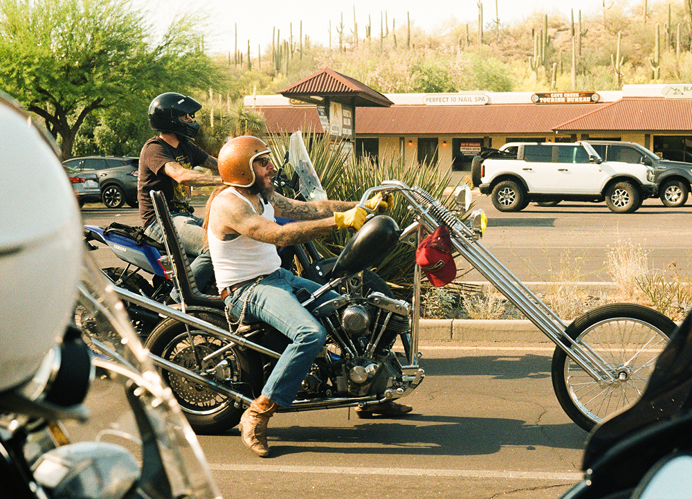 Custom chopper motorcycle ridden through city street, long front fork, helmeted riders, storefronts behind.