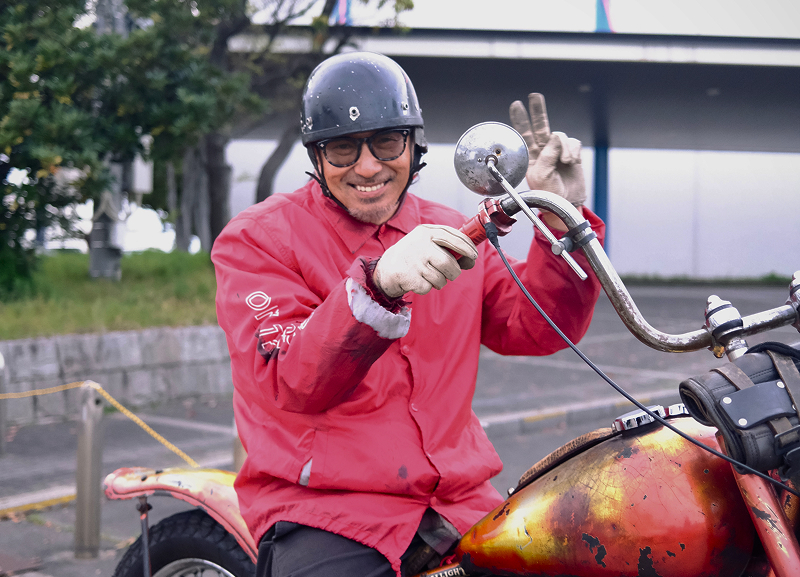 Rider on a red custom chopper flashing peace sign toward camera while stopped with buildings behind