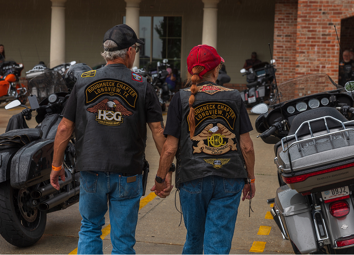 A couple holds hands and walks away from the camera, wearing vests with Harley Owner’s Group patches