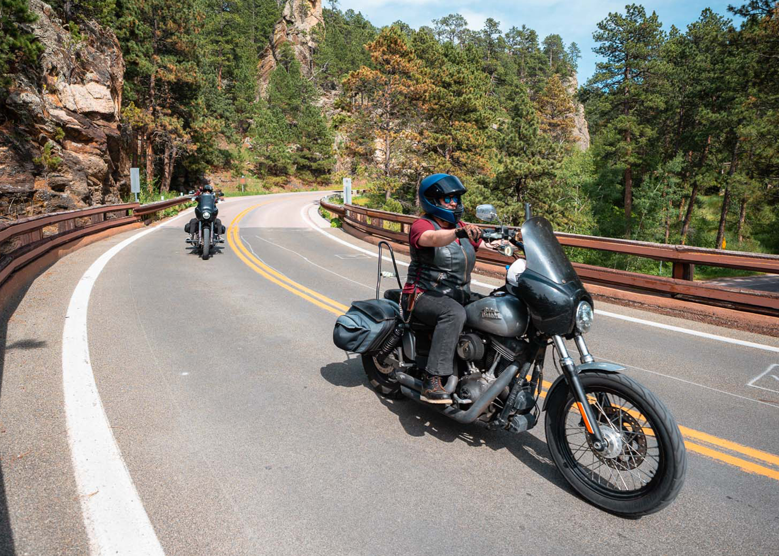Two motorcyclists ride through a forested curve on a mountain road lined with guardrails and rocky cliffs.