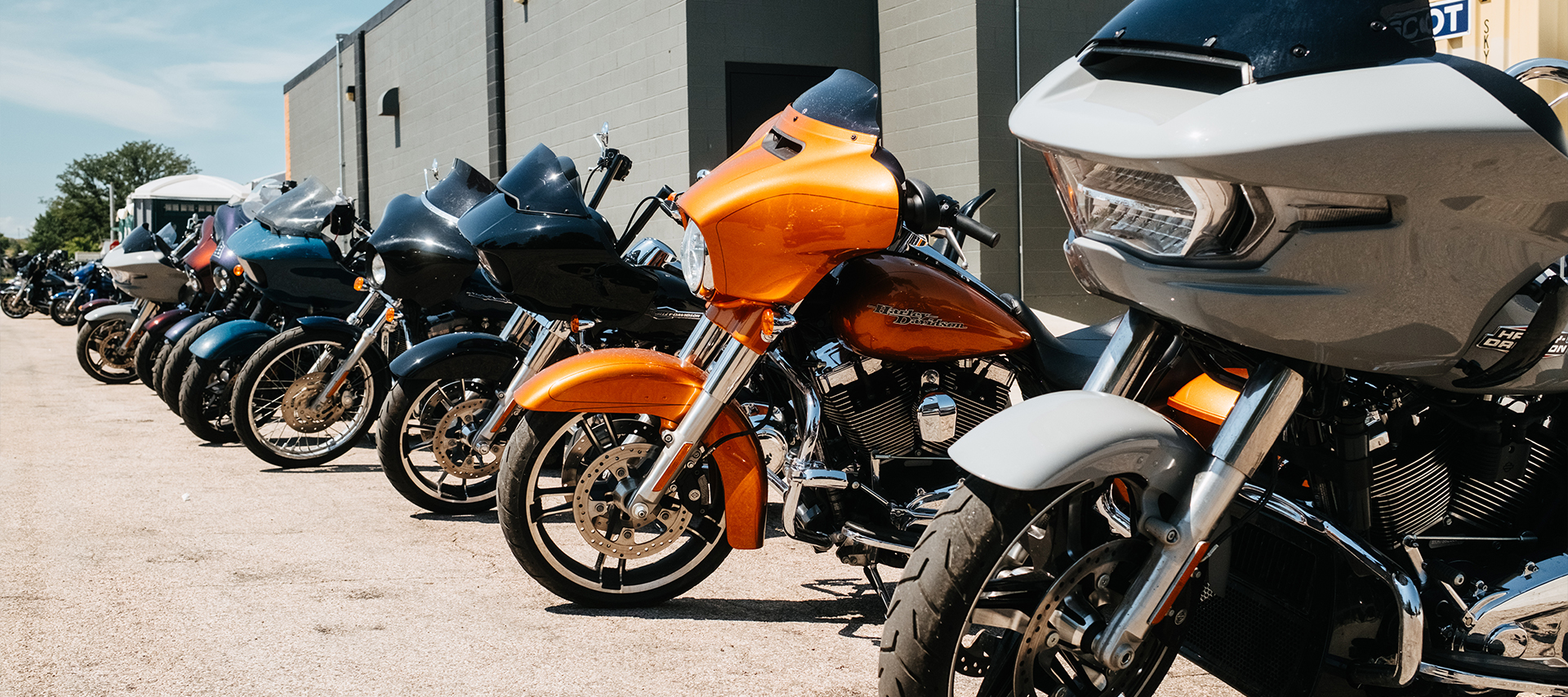 A long row of Harley-Davidson motorcycles parked outside the Sturgis Harley-Davidson dealership, featuring a striking orange touring bike in front.