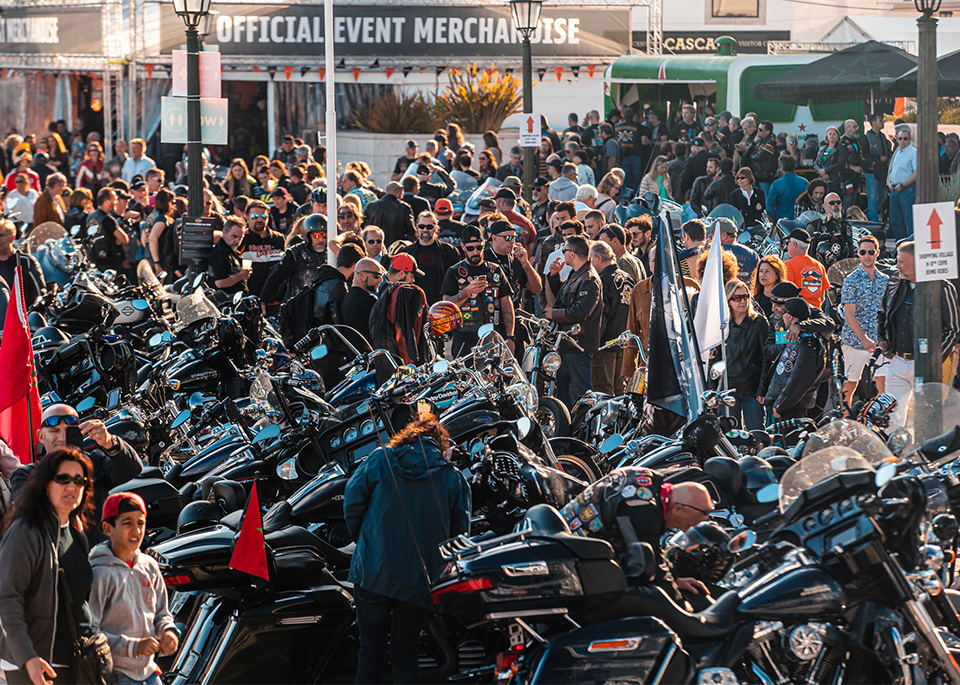 Foule se rassemblant autour de motos garées lors d’un événement en extérieur, des tentes d'exposants apparaissant en arrière-plan