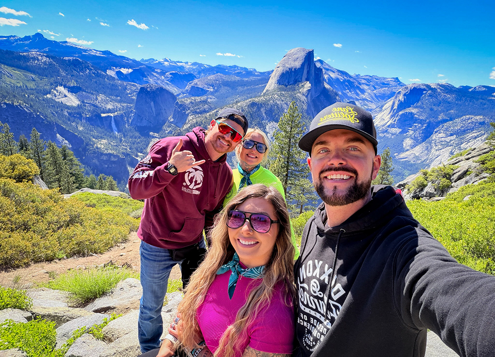 Four travelers take a selfie at a Yosemite viewpoint with Half Dome and mountain scenery behind them