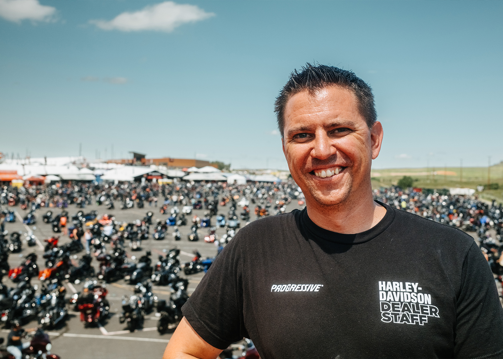 Smiling Black Hills Harley-Davidson staff member stands in front of a massive motorcycle rally crowd outdoors.