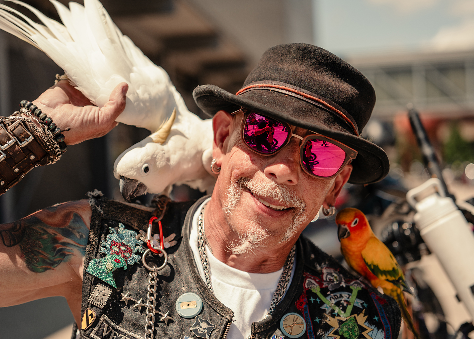 An amusing older man wearing a bikers vest and pink glasses with a cockatoo on his shoulder