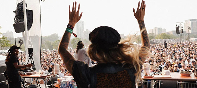 A woman from behind raising her hands in the air on stage in front of a large crowd.
