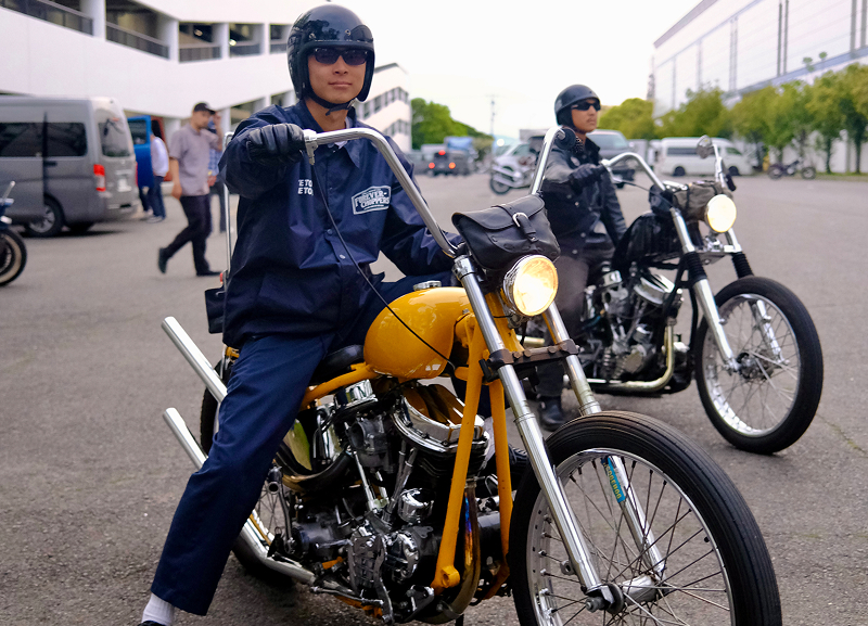 Rider seated on a gold custom motorcycle parked in an open lot with another rider and bike nearby