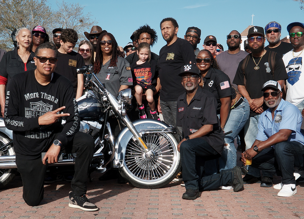 Group gathered outdoors around a classic Harley-Davidson motorcycle for a photo, wearing branded shirts and caps on a sunny day