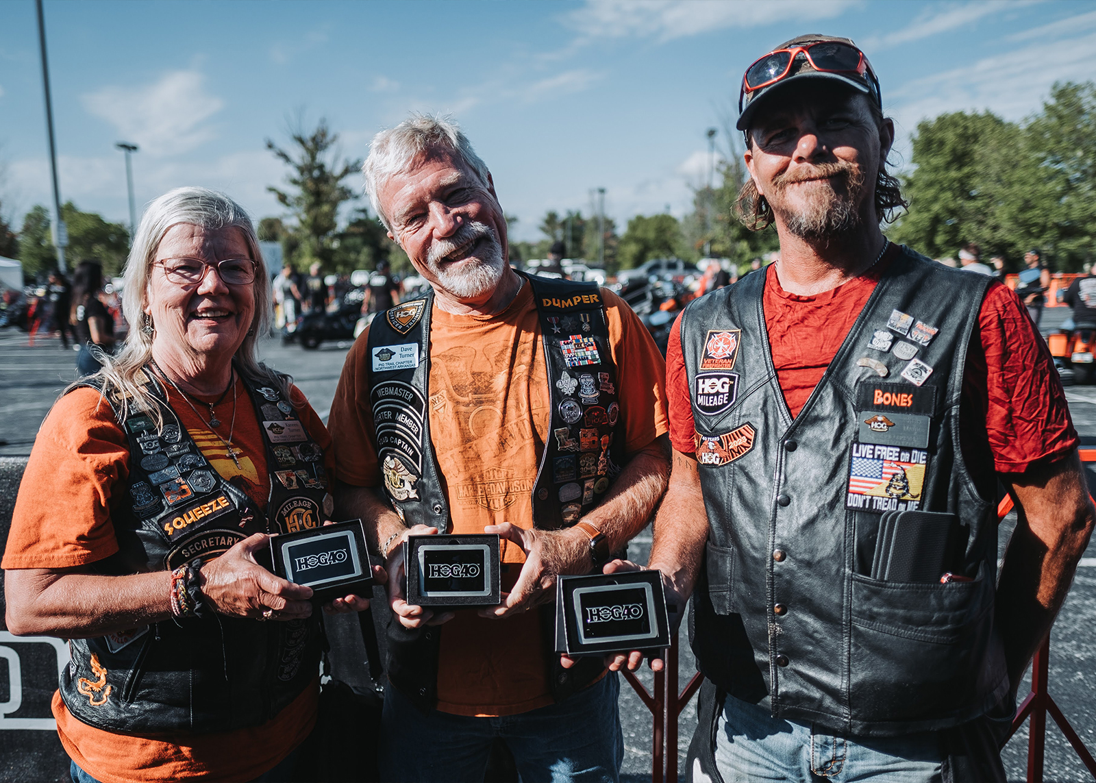 Three H.O.G. members pose wearing leather vests covered in patches and holding souvenirs that say HOG40 on them
