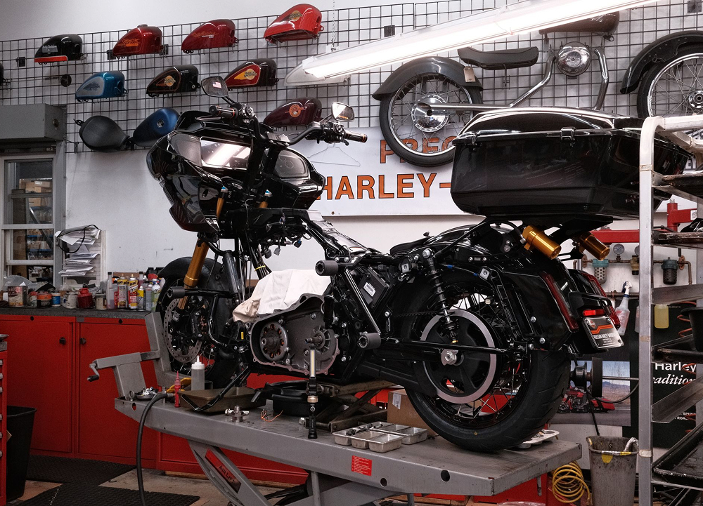 Disassembled H-D motorcycle on lift in service bay with exposed engine parts, tools, and tanks on wall behind