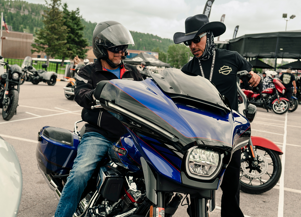 Person seated on blue H-D motorcycle while staff member explains controls in lot with other bikes and tents in background.