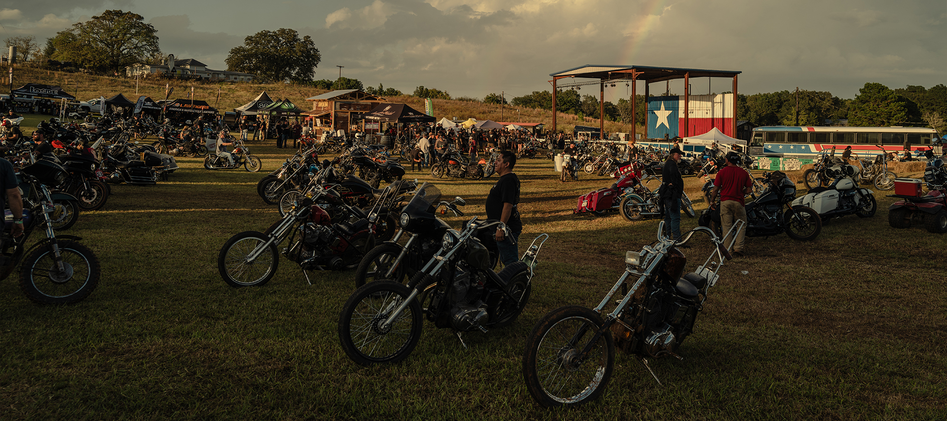 People look at rows of show bikes that are parked on a large grass field with a stage, Texas flag, and rainbow in the background.