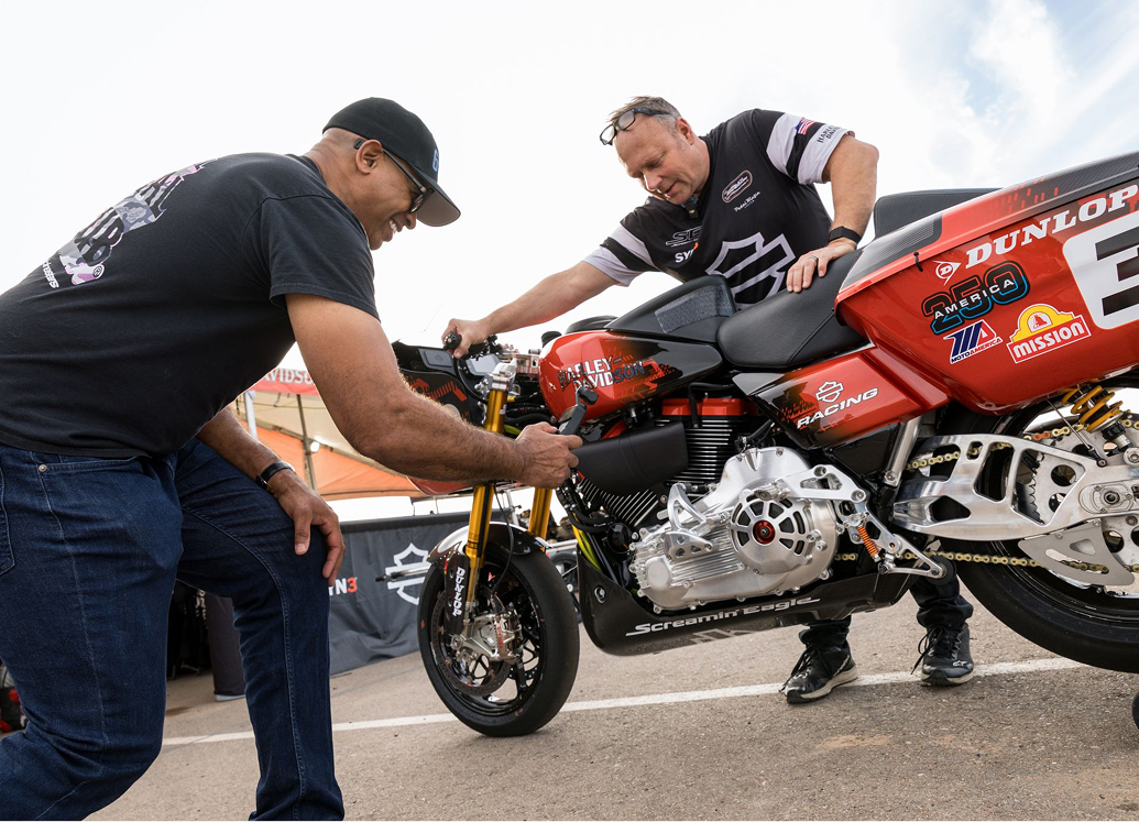 The author takes a close up recording of a race motorcycle as a crew member wheels it to the garage