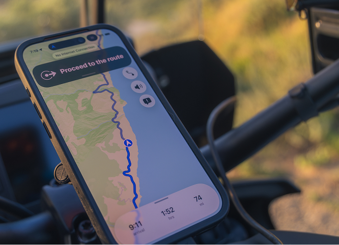 Smartphone mounted on motorcycle handlebars displays a navigation route along a winding coastal road with distance and time details visible