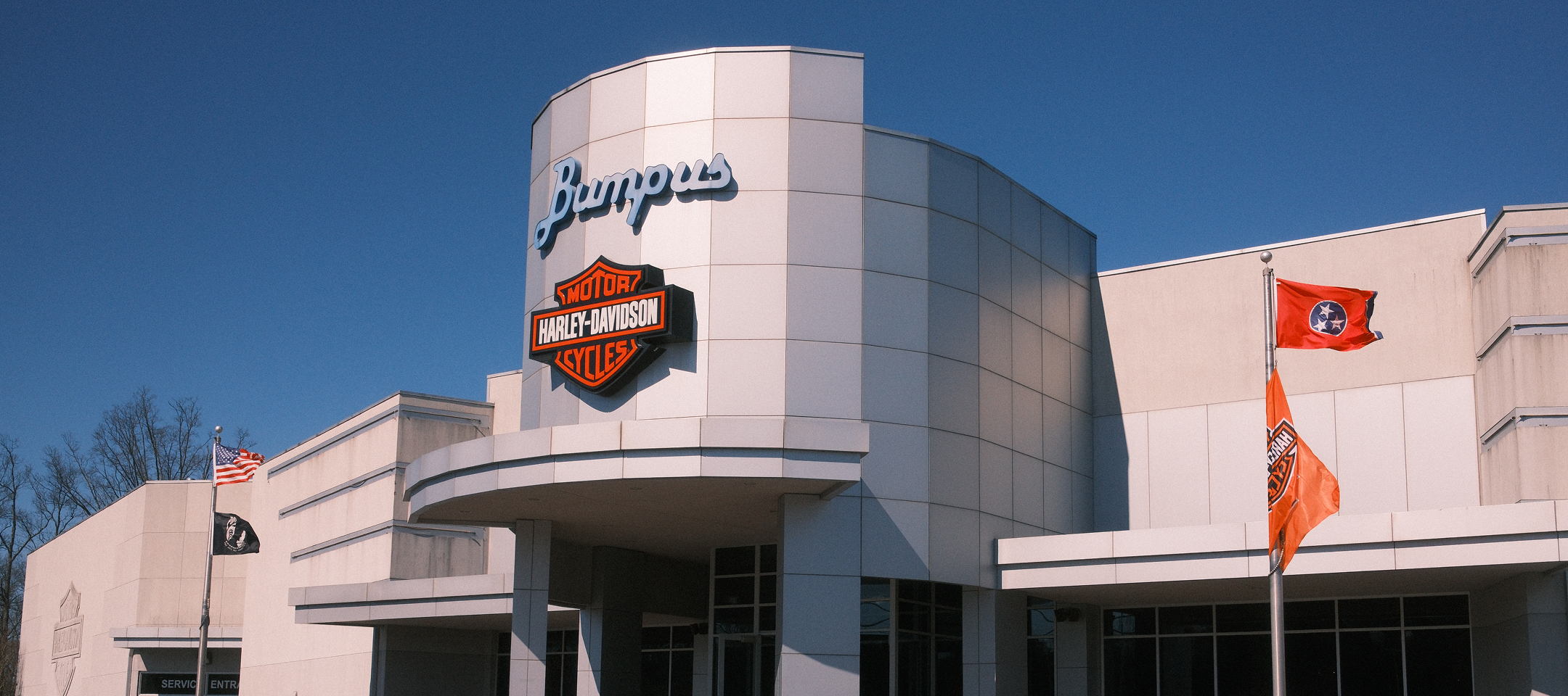 Exterior of Bumpus Harley-Davidson dealership with modern facade, logo signage, and flags under blue sky
