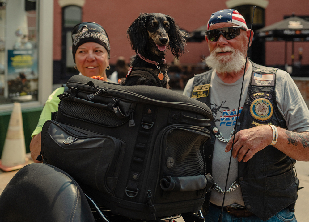 Two travelers pose with a dog riding in a motorcycle luggage rack during a downtown rally gathering
