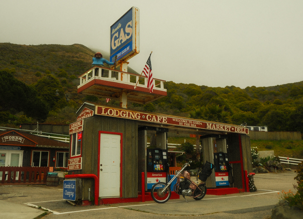 Roadside lodging, café, and gas station with vintage signage, custom motorcycle, and coastal hills