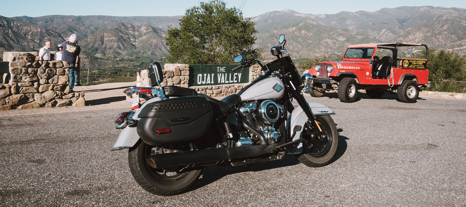 A Harley-Davidson Heritage Classic motorcycle parked at Ojai Valley overlook with mountains and a classic red Jeep in the background