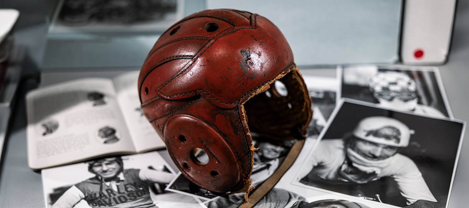 A vintage leather motorcycle helmet with a wing shape on the front is displayed on a table surrounded by black and white archival photos of riders.