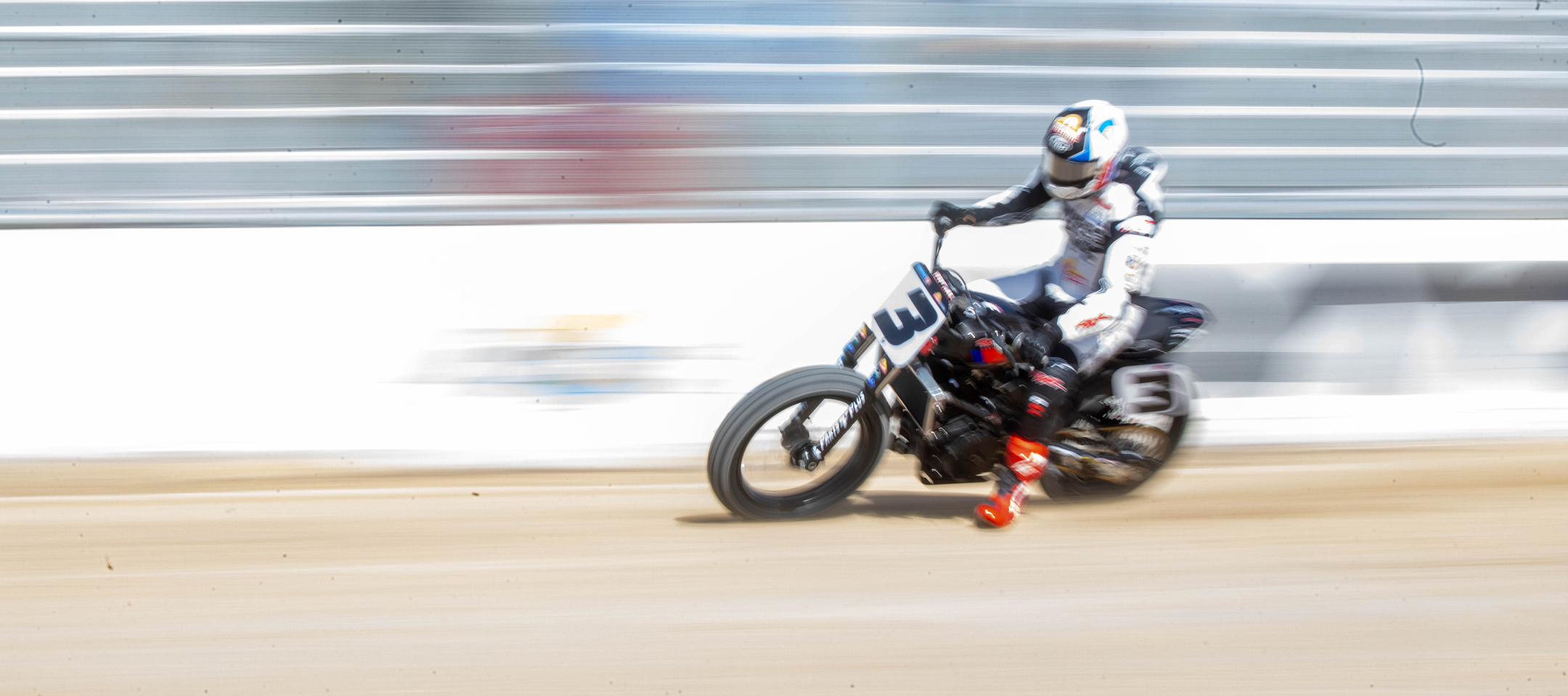 Flat track rider speeding through dirt straightaway with motion blur trailing behind and bright red boots skimming track surface