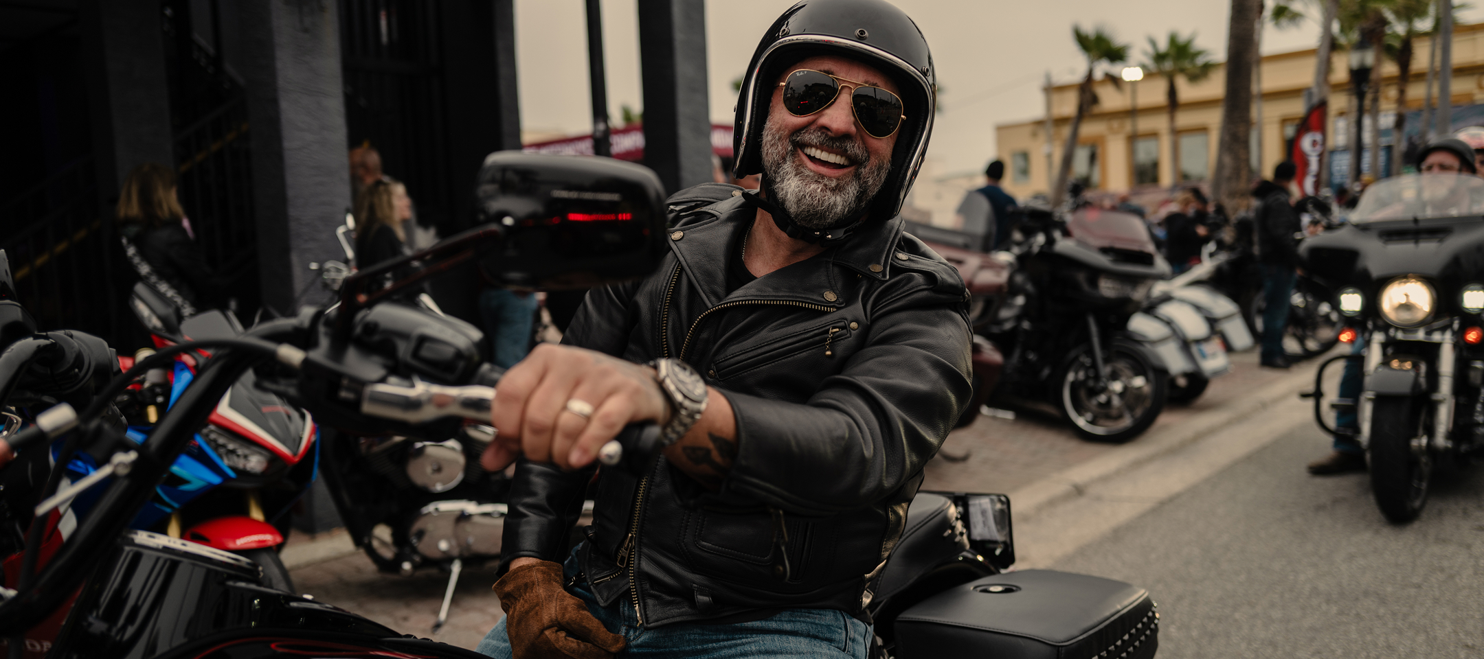 Motorcyclist smiles at the camera, wearing a leather jacket and helmet, with parked motorcycles lining the street