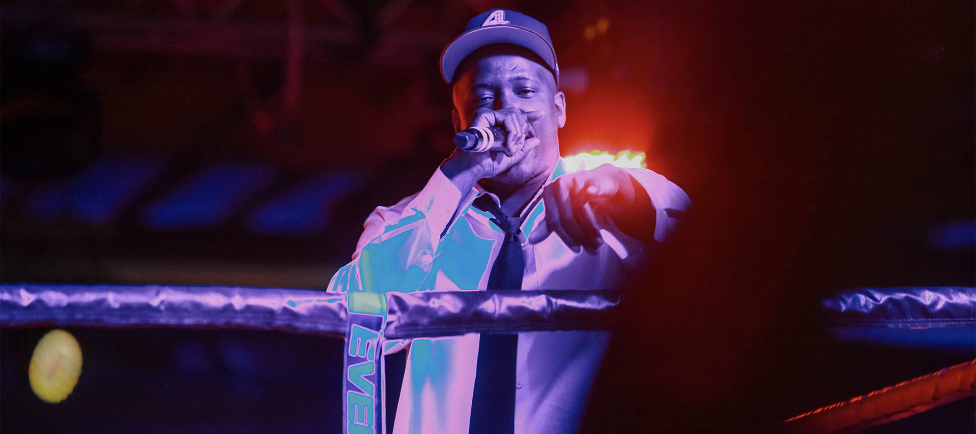 Rapper YG in a white long sleeve shirt, black tie and baseball hat looks and points at the camera while performing from the inside of a boxing ring.