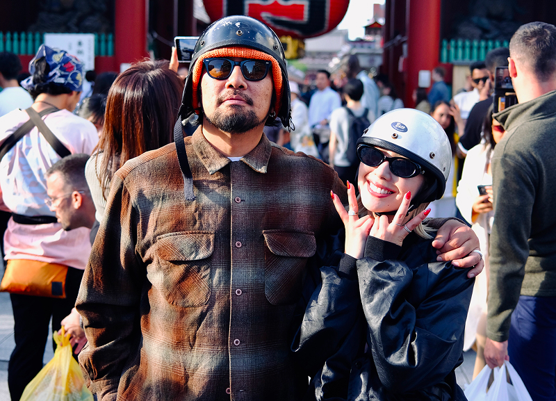 Yusuke and Shino wearing helmets standing in front of a crowd with a large lantern in the background