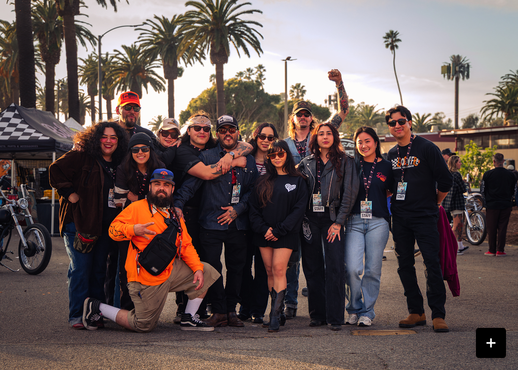 Group of people posing together at an outdoor motorcycle event lined with palm trees