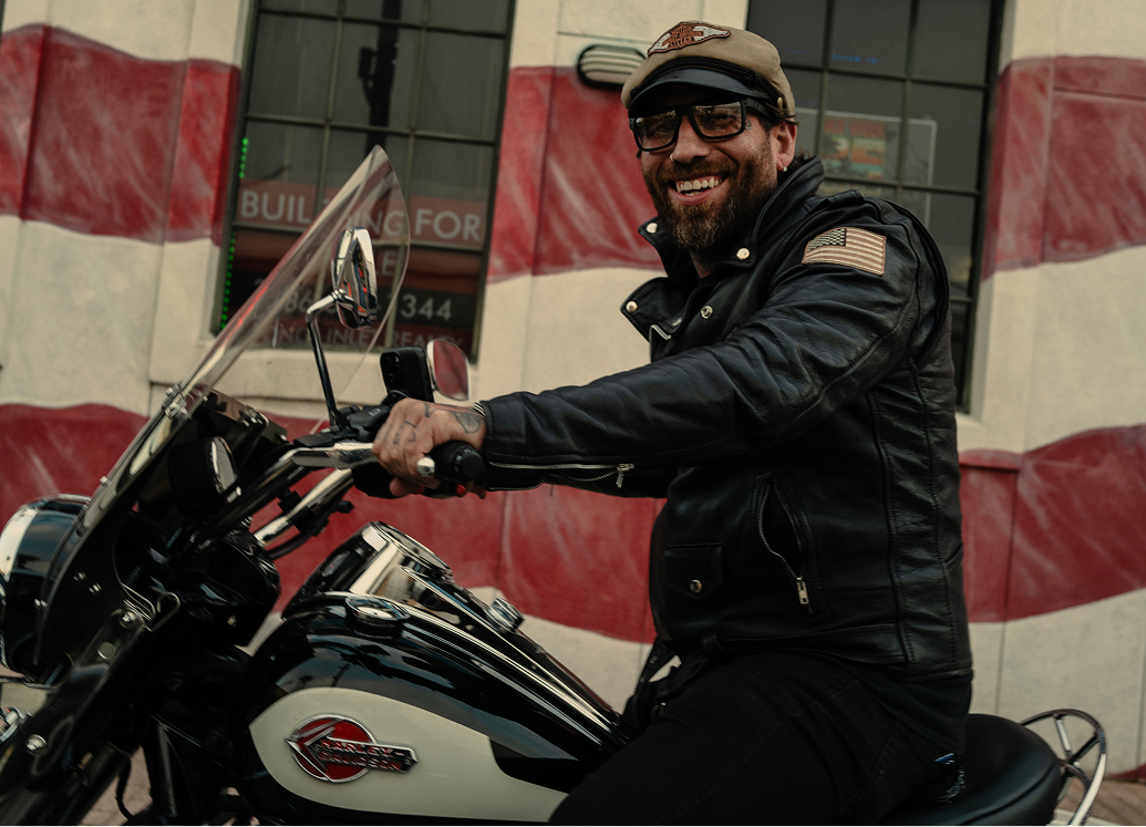 Rider seated on a classic motorcycle in front of a large American flag mural at Daytona Bike Week