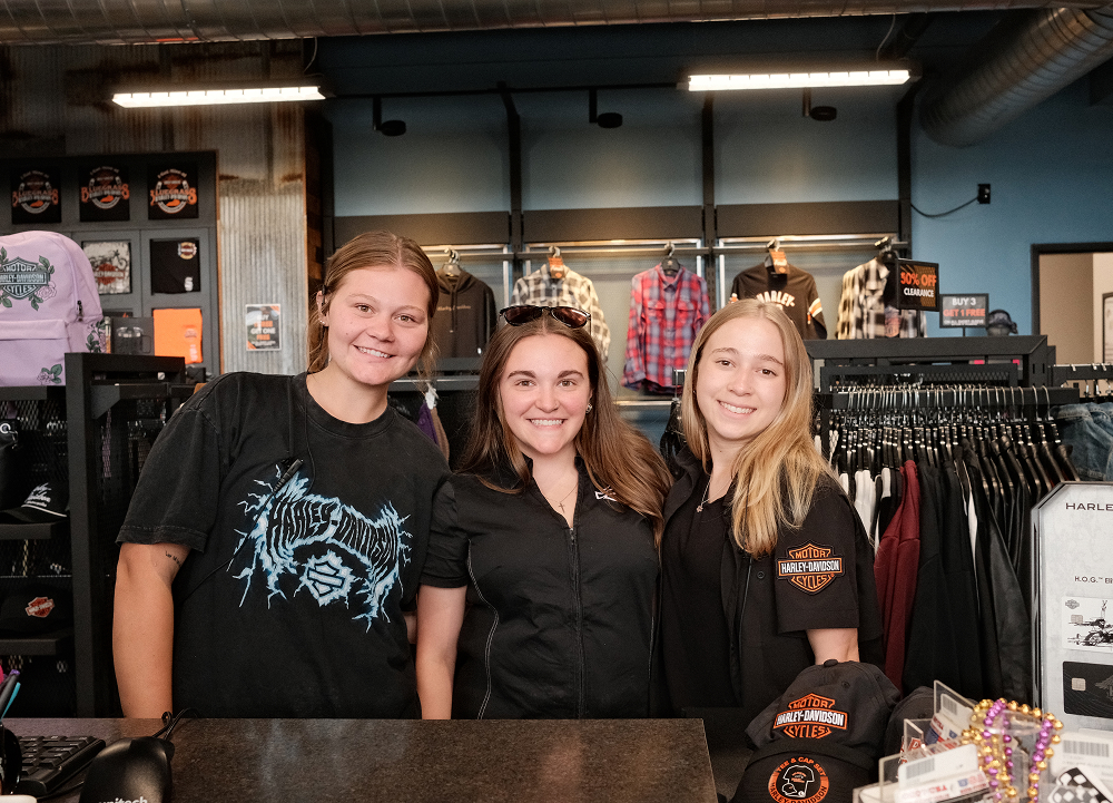 Three dealership team members standing behind a Harley-Davidson apparel counter surrounded by clothing displays