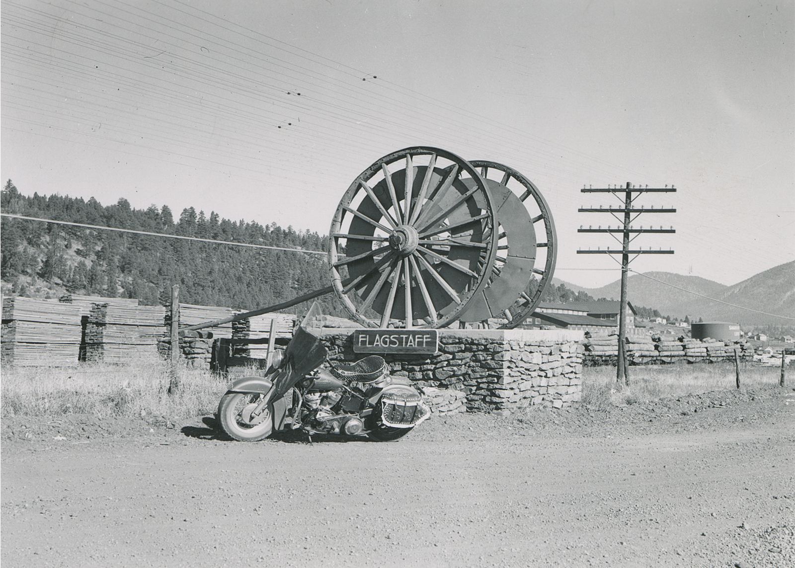 Black and white historical photo of Eldred Harrington’s 1951 Harley-Davidson Hydra Glide motorcycle parked by a large wooden wheel with a "Flagstaff" sign in Arizona.