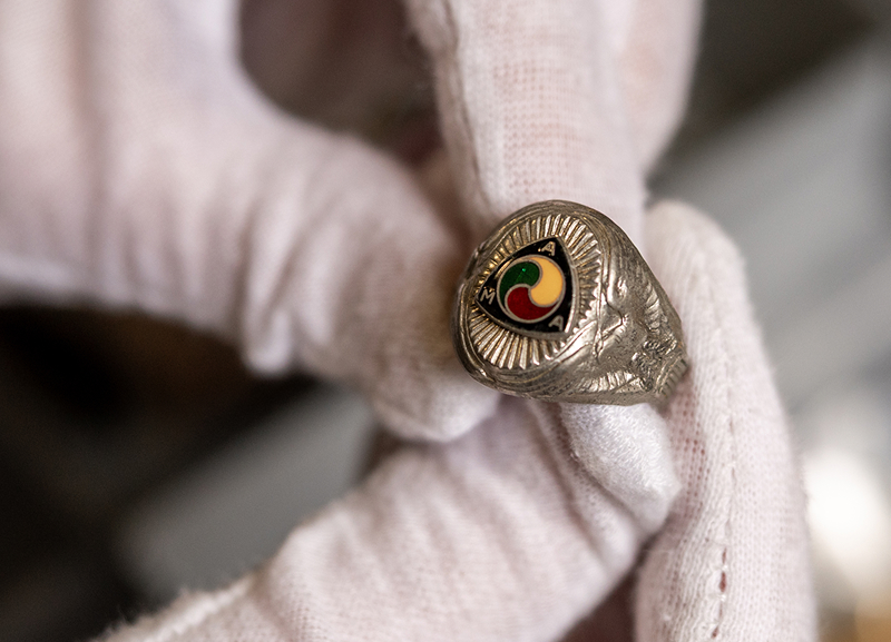 Gloved hands holding a ring with a small AMA emblem face, displayed above a drawer of vintage items
