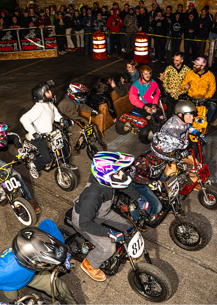 Riders on small motorcycles line up on an indoor track surrounded by spectators and barriers