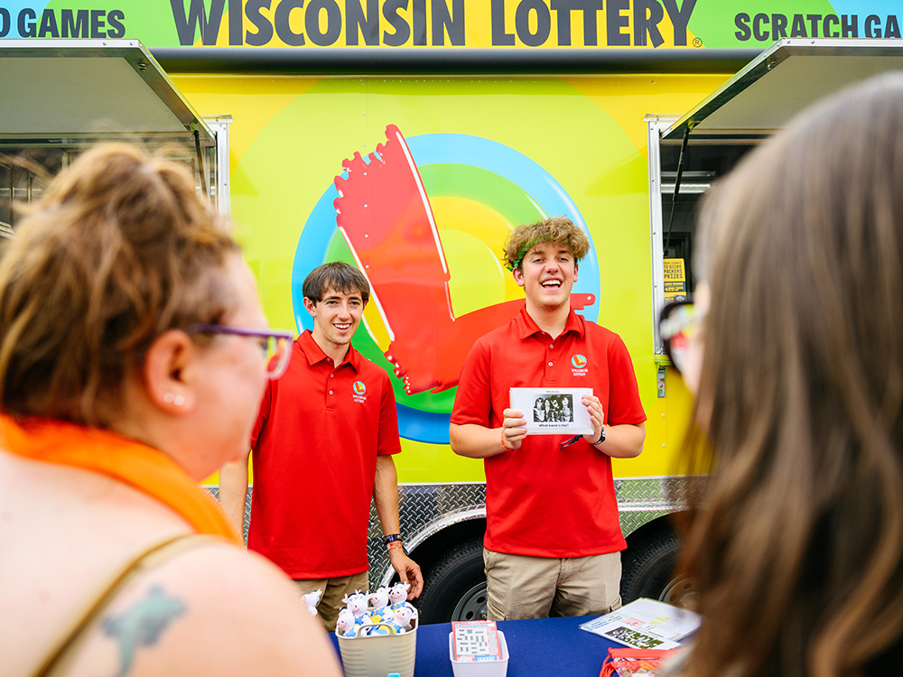 Personal del stand de la Lotería de Wisconsin con camisetas rojas repartiendo tarjetas rasca y gana