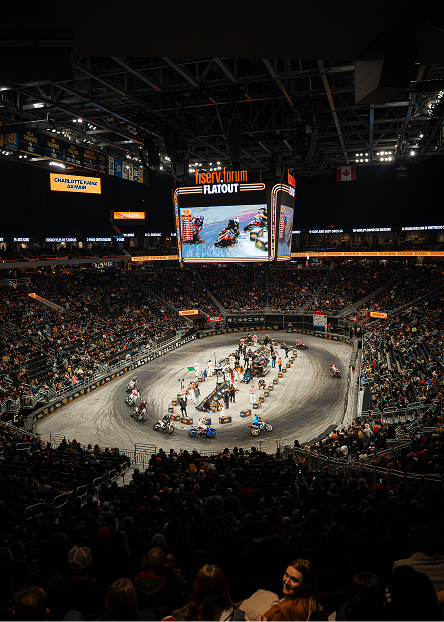 Large indoor arena packed with spectators watching multiple riders race around the dirt track