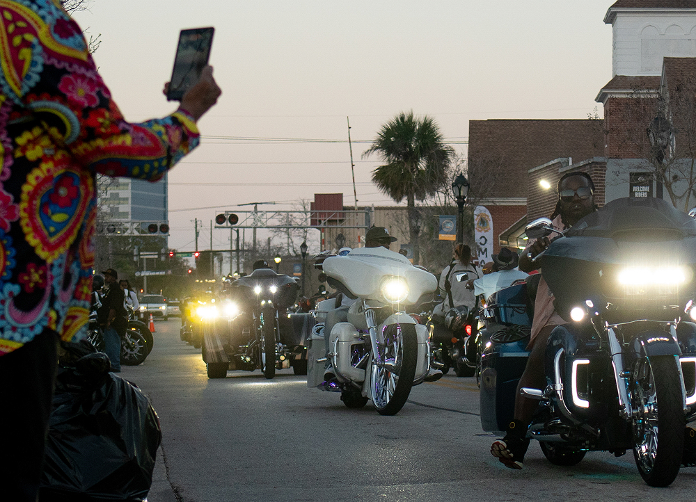 Motorcycles ride down a city street at dusk with headlights on as a person records the procession