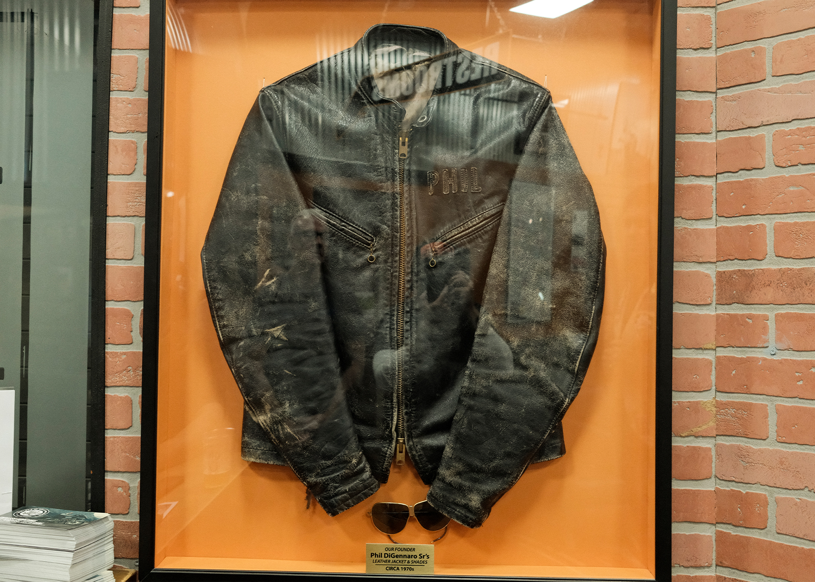 Phil DiGennaro Sr.’s worn black leather jacket displayed in a glass frame against an orange background.