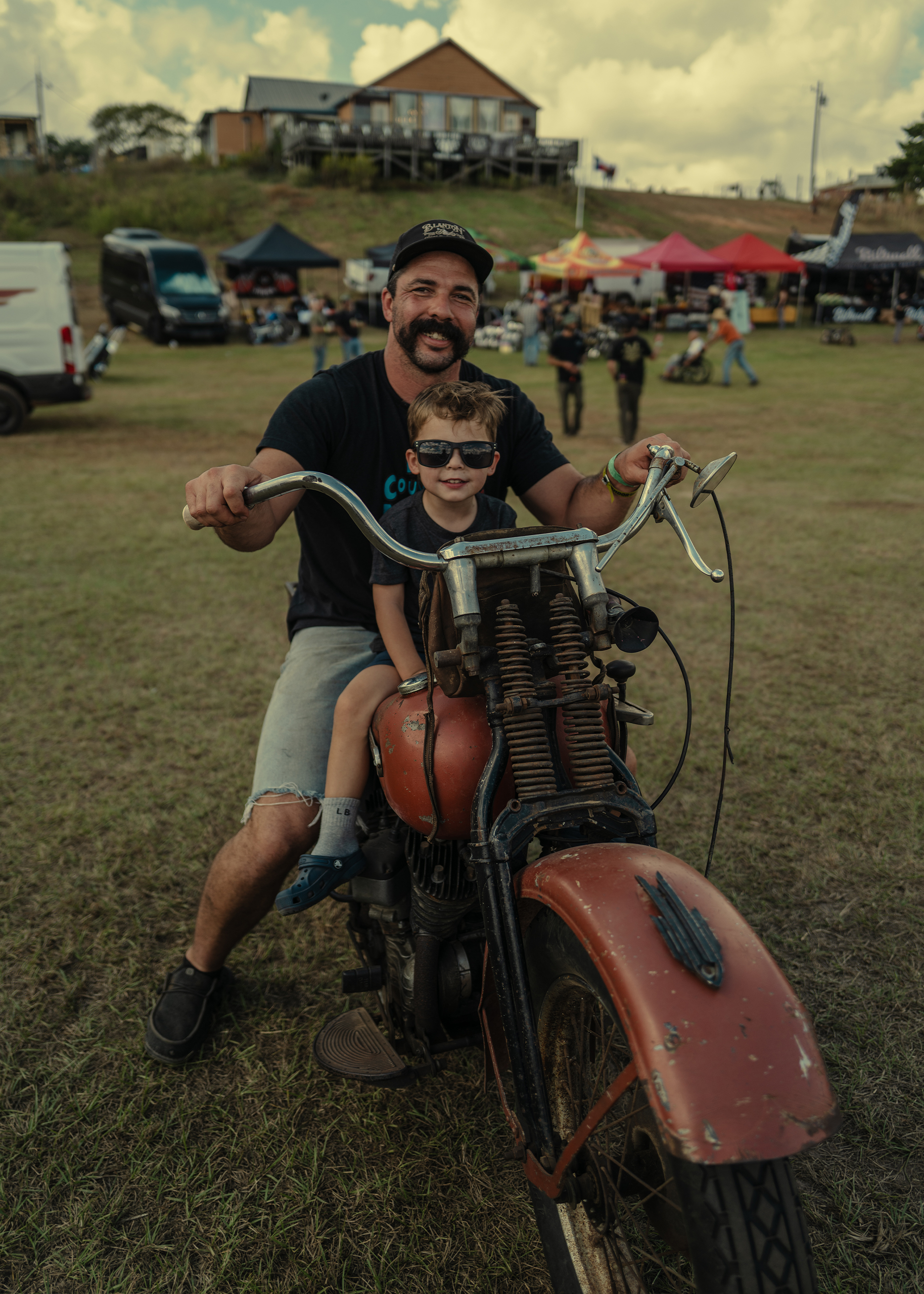A father and son smile while sitting together on a vintage Harley-Davidson motorcycle at the Born Free Texas rally.