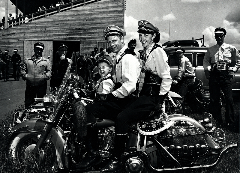 Archival photograph of riders seated on a decorated motorcycle surrounded by spectators at an event