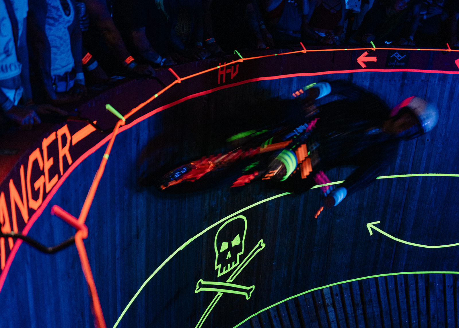 A motorcyclist rides the Wall of Death under blacklight, surrounded by neon arrows and warning signs.