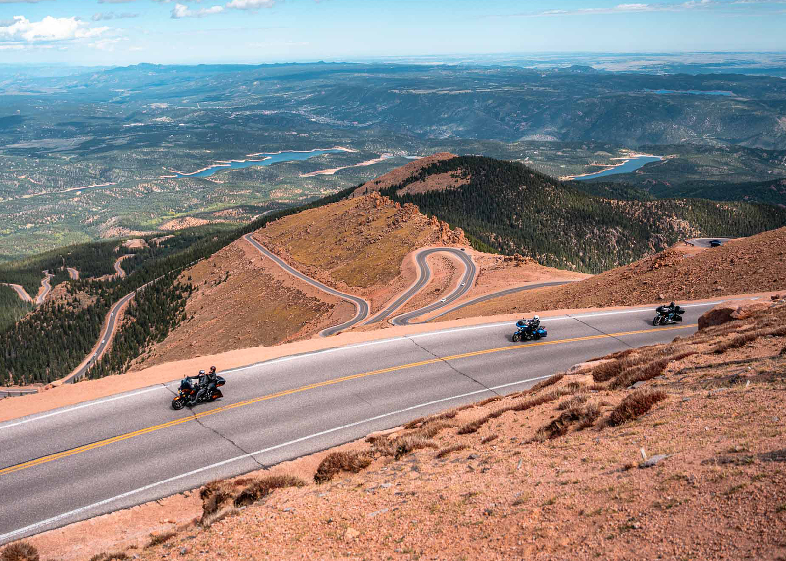 A wide shot looking down of three Harley-Davidson motorcycles ascending Pikes Peak, with the road winding in the distance and landscape extending to the horizon.
