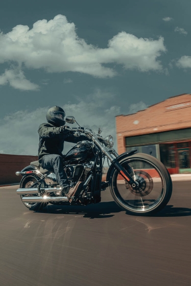 A motorcycle rider cruises through a small town on a 2026 H-D Breakout motorcycle as they pass by brick and storefront buildings.