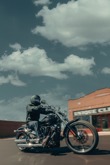 A motorcycle rider cruises through a small town on a 2026 H-D Breakout motorcycle as they pass by brick and storefront buildings.